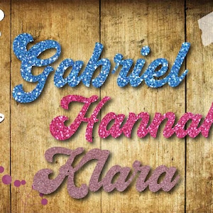 Könnte beinhalten: Drei Namen, Gabriel, Hannah und Klara, sind in glitzernden Buchstaben auf einem Holzgrund geschrieben. Die Buchstaben sind blau, rosa und lila.