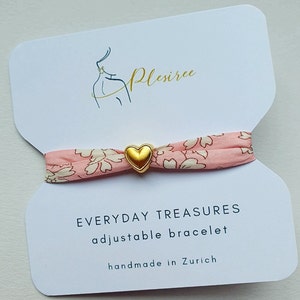 May include: A pink and white floral patterned fabric bracelet with a gold heart charm. The bracelet is adjustable and says "Everyday Treasures adjustable bracelet handmade in Zurich".