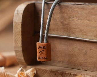 Könnte beinhalten: Ein hölzernes Baby-Andenken mit dem Namen "Lukas", Fußabdrücken und dem Datum "26.05.24" eingraviert. Es ist an einem grauen Seil befestigt. Im Hintergrund eine Holzkiste und Holzspäne.