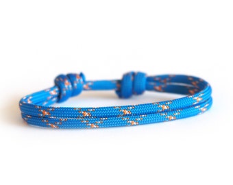 Può includere: Un braccialetto di corda blu con dettagli arancioni e bianchi. Il braccialetto è annodato alle due estremità ed è realizzato in un materiale strutturato e resistente. Il braccialetto è progettato per essere indossato al polso.