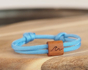 May include: A light blue bracelet with a wooden accent. The bracelet is made of two strands of woven cord, secured with knots. The wooden accent features a carved wave design. The bracelet is displayed on a wooden surface.