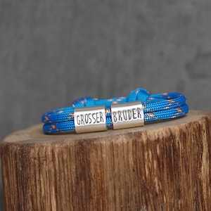 May include: A blue and orange rope bracelet with the words "GROSSER BRUDER" engraved on silver metal clasps. The bracelet is resting on a wooden surface against a gray background. The bracelet is a unique accessory.