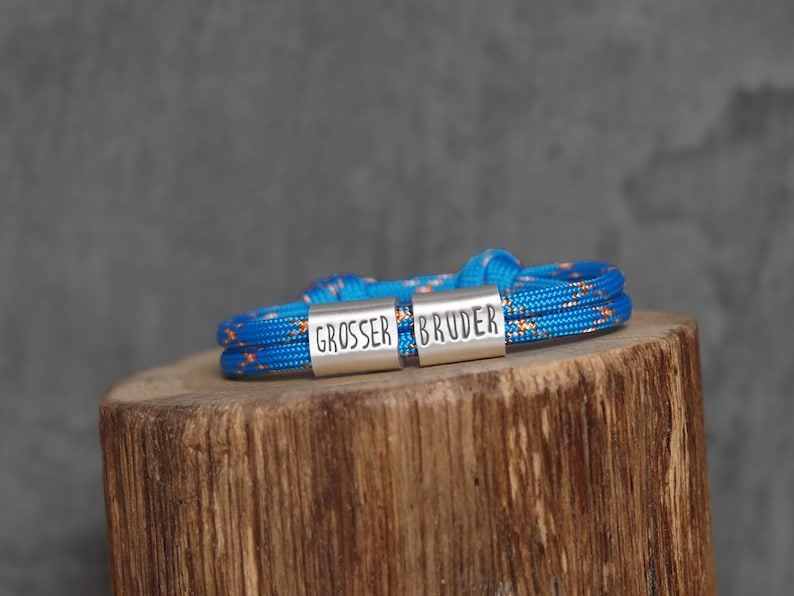 May include: A blue and orange rope bracelet with the words "GROSSER BRUDER" engraved on silver metal clasps. The bracelet is resting on a wooden surface against a gray background. The bracelet is a unique accessory.