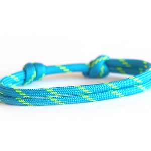 Op de afbeelding: Een turquoise en gele touw armband. De armband is gemaakt van een dubbele streng turquoise touw met een geel patroon. De armband is vastgebonden met twee knopen, één aan elk uiteinde. De achtergrond is wit.