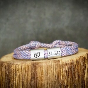 May include: A lavender and white rope bracelet with two silver-colored rectangular clasps. One clasp features two baby footprints, and the other has the date "31.5.20" engraved. The bracelet is resting on a wooden surface.