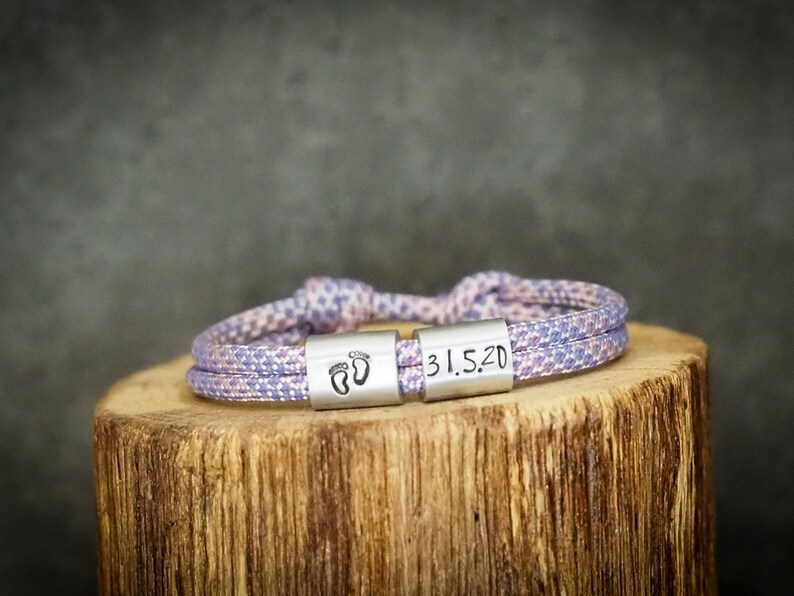 May include: A lavender and white rope bracelet with two silver-colored rectangular clasps. One clasp features two baby footprints, and the other has the date "31.5.20" engraved. The bracelet is resting on a wooden surface.