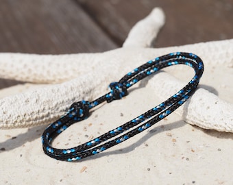 Può includere: Un braccialetto in corda nero e blu a tema nautico. Il braccialetto presenta un design intrecciato con accenti blu e bianchi. Il braccialetto è appoggiato su una superficie sabbiosa con una stella marina bianca sullo sfondo.