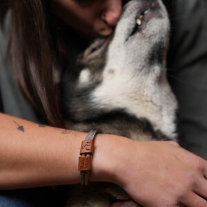 Könnte beinhalten: Ein silberfarbenes Armband mit zwei rechteckigen braunen Lederakzenten. Die Lederstücke sind mit den Worten "Balou Milow" graviert. Das Armband befindet sich am Handgelenk einer Person, mit einem Hund im Hintergrund.