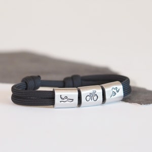 Può includere: Un braccialetto con cordino nero con tre perline cilindriche color argento. Ogni perlina presenta un'icona incisa diversa: un nuotatore, una bicicletta e un corridore. Il braccialetto è progettato per atleti e appassionati di sport.