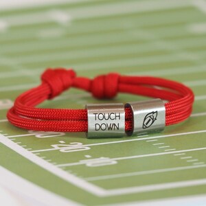 Könnte beinhalten: Rotes Seilarmband mit zwei silberfarbenen Metallperlen. Eine Perle trägt die Aufschrift "TOUCH DOWN", die andere ein Football-Motiv. Das Armband wird auf einem grünen Hintergrund mit Footballfeldmarkierungen präsentiert.