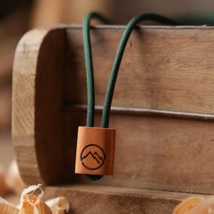 Puede incluir: Un collar con colgante de madera con un cordón verde oscuro. El colgante es un pequeño bloque rectangular de madera con un diseño de cordillera grabado. El collar se exhibe sobre una superficie de madera.