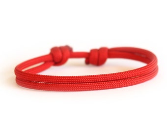 Könnte beinhalten: Ein leuchtend rotes Armband aus geflochtenem Kordel. Das Armband hat zwei parallele Kordelstränge, die an jedem Ende durch Knoten verbunden sind. Das verstellbare Design ermöglicht eine bequeme Passform. Dieses Accessoire ist für den Alltag geeignet.