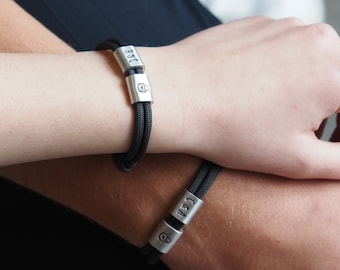 May include: Two black rope bracelets with silver-colored clasps. Each clasp has initials and a small symbol. The bracelets are worn on wrists, suggesting a matching set or a couple's accessory.