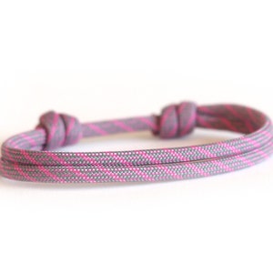 Könnte beinhalten: Ein graues und rosa Seilarmband mit zwei Knoten. Das Armband besteht aus einem strapazierfähigen, gewebten Material. Das Farbschema ist eine Kombination aus Grau und Rosa und schafft ein modernes und stilvolles Accessoire.