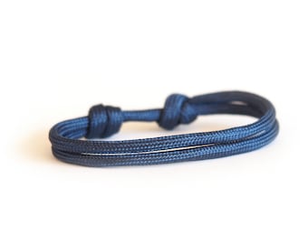 Può includere: Un braccialetto in corda blu navy con due fili. Il braccialetto è annodato ad ogni estremità e al centro, creando un design semplice ma elegante. Il braccialetto è posto su uno sfondo bianco.
