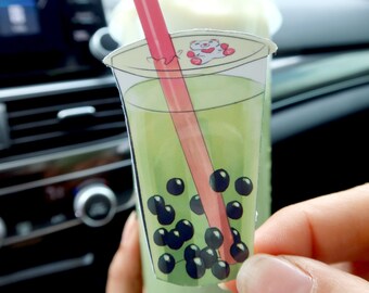 May include: A green tea boba drink with tapioca pearls and a pink straw in a clear plastic cup. The cup has a white lid with a cartoon bear on it.
