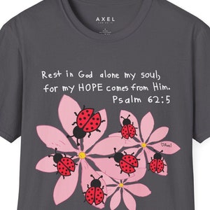 Pode incluir: Uma camiseta cinza escura com um design de flores rosa e joaninhas vermelhas. A camiseta tem o texto "Rest in God alone my soul, for my HOPE comes from Him. Psalm 62:5" impresso nela.