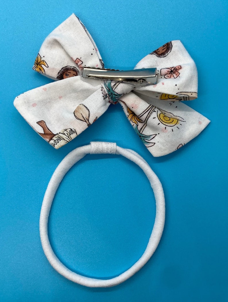 Pode incluir: Um la&ccedil;o de tecido branco com um clipe prateado e uma faixa el&aacute;stica branca. O la&ccedil;o tem uma estampa de uma menina de desenho animado com flores e um sol.