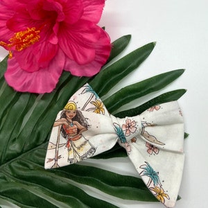 Pode incluir: Um la&ccedil;o branco com um desenho estampado de um personagem de desenho animado com cabelo castanho, um chap&eacute;u amarelo e um colar azul. O personagem est&aacute; em p&eacute; numa praia com palmeiras e flores. O la&ccedil;o est&aacute; sobre uma folha verde.