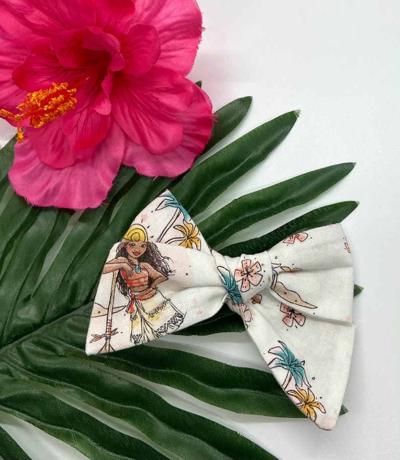 Pode incluir: Um la&ccedil;o branco com um desenho estampado de um personagem de desenho animado com cabelo castanho, um chap&eacute;u amarelo e um colar azul. O personagem est&aacute; em p&eacute; numa praia com palmeiras e flores. O la&ccedil;o est&aacute; sobre uma folha verde.