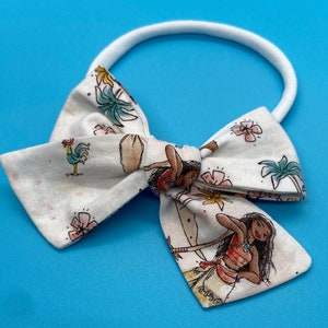 Pode incluir: Um la&ccedil;o de tecido branco com uma estampa de Moana que apresenta palmeiras, flores e Moana. O la&ccedil;o est&aacute; preso a uma faixa el&aacute;stica branca.