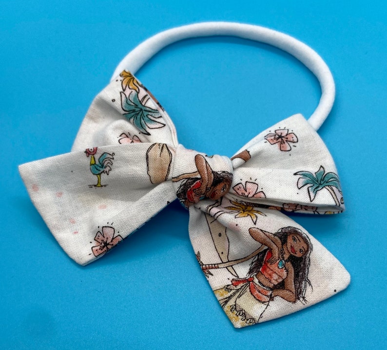 Pode incluir: Um la&ccedil;o de tecido branco com uma estampa de Moana que apresenta palmeiras, flores e Moana. O la&ccedil;o est&aacute; preso a uma faixa el&aacute;stica branca.