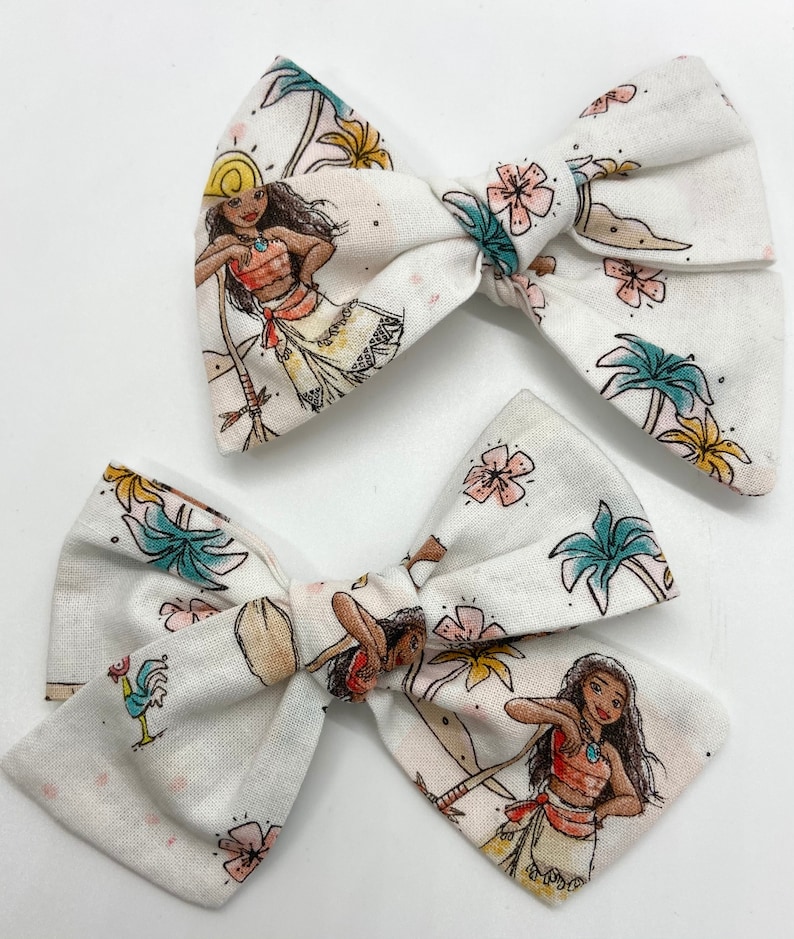 Pode incluir: Dois la&ccedil;os de cabelo de tecido branco com uma estampa tropical que apresenta um personagem de desenho animado. A estampa inclui palmeiras, flores e um galo.