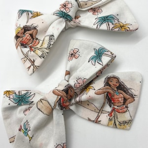 Pode incluir: Dois la&ccedil;os de cabelo de tecido branco com uma estampa de uma mulher polin&eacute;sia de desenho animado em um ambiente tropical. A estampa apresenta palmeiras, flores e um oceano azul.