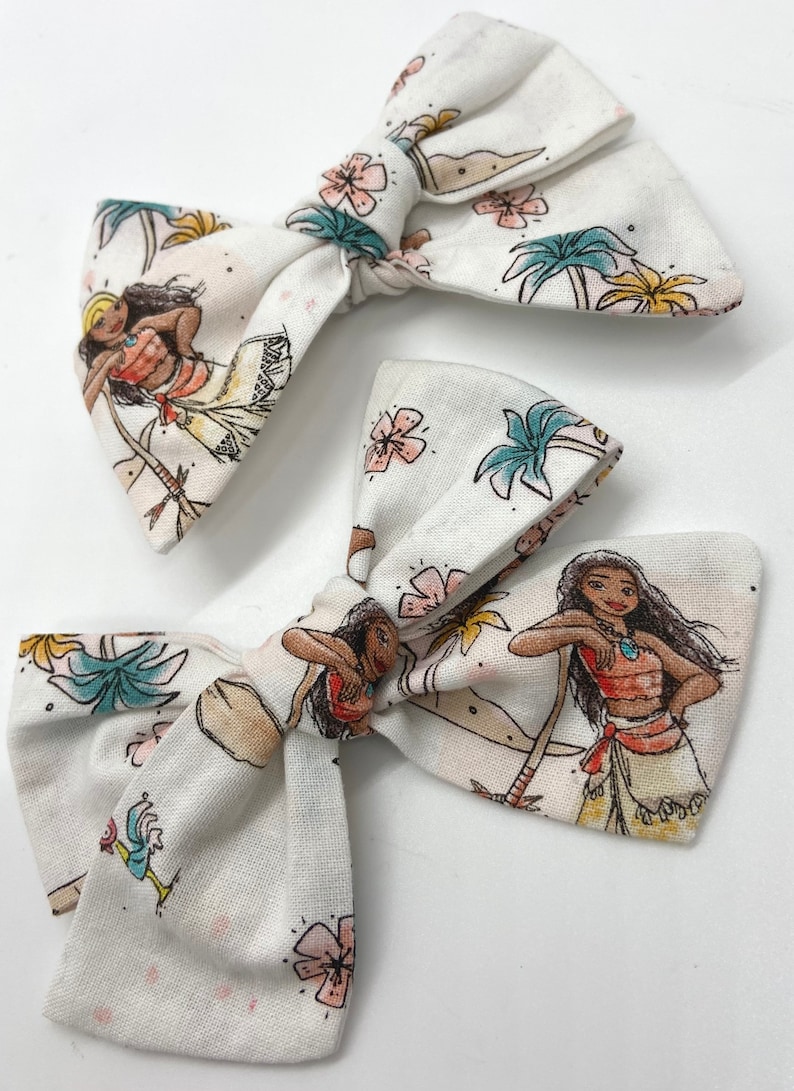 Pode incluir: Dois la&ccedil;os de cabelo de tecido branco com uma estampa de uma mulher polin&eacute;sia de desenho animado em um ambiente tropical. A estampa apresenta palmeiras, flores e um oceano azul.