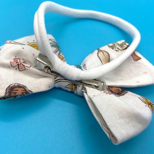 Pode incluir: Faixa de cabelo de tecido branco com um la&ccedil;o com estampa de meninas e flores coloridas. A faixa tem uma banda el&aacute;stica branca e um clipe prateado.