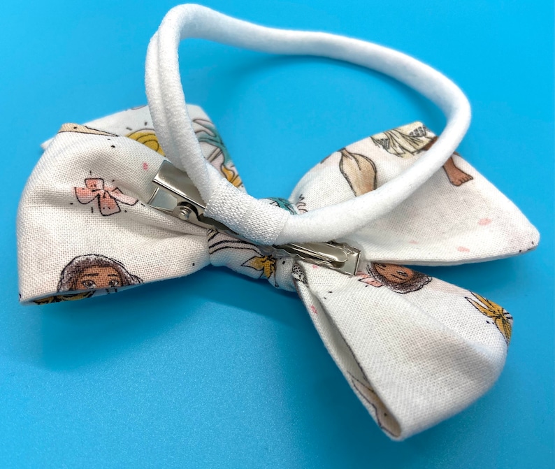 Pode incluir: Faixa de cabelo de tecido branco com um la&ccedil;o com estampa de meninas e flores coloridas. A faixa tem uma banda el&aacute;stica branca e um clipe prateado.