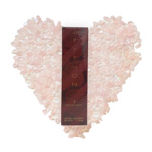 Puede incluir: Un frasco rojo y dorado de desodorante Trussardi Python Sensual. El frasco está rodeado de piedras de cuarzo rosa en forma de corazón.