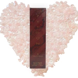 Puede incluir: Caja de perfume rectangular burdeos con la palabra "PYTHON" en letras doradas. La caja tiene un patrón de piel de serpiente. La caja está rodeada de piedras rosas en forma de corazón. El perfume es de 50 ml, 1,7 fl oz.