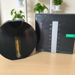 Pode incluir: Um frasco de perfume preto com um atomizador dourado está ao lado de uma caixa preta. A caixa tem as palavras "man inferno paradiso" e "United Colors of Benetton". O frasco contém 50 ml.