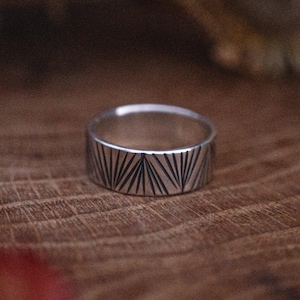 Könnte beinhalten: Ein silberner Ring mit geometrischem Muster. Der Ring hat geätzte Linien, die ein fächerartiges Muster um das Band bilden. Er wird auf einer Holzoberfläche mit getrockneten Blumen im Hintergrund präsentiert.