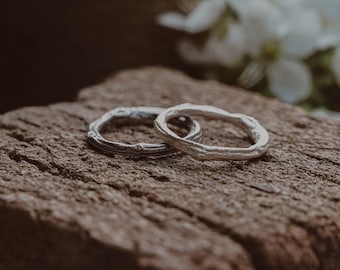 May include: Two silver wedding bands with a unique, organic, twig-like design, resting on a rough, brown stone surface.