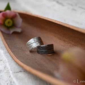 Könnte beinhalten: Zwei Ringe mit Blattstruktur, einer silberfarben und einer dunkelgrau, liegen in einer Holzschale mit einer Blume. Die Ringe haben ein detailliertes Blättermuster. Die Schale hat eine warme braune Farbe.