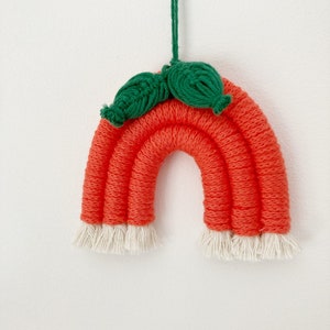 May include: A handmade orange and white rainbow wall hanging with green leaves at the top. The rainbow is made of yarn and has a fringe at the bottom.