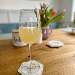 Könnte beinhalten: Ein klares Glas mit einer personalisierten Inschrift "Mama", gefüllt mit einem hellgelben Getränk. Das Glas steht auf einem weißen Untersetzer mit den Worten "Gonn' glas" in goldener Schrift.