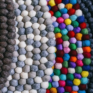 May include: A close-up of a colorful rug made of felt balls. The rug is made up of four sections, each with a different color scheme. The first section is black, the second is gray and white, the third is a rainbow of colors, and the fourth is dark blue.