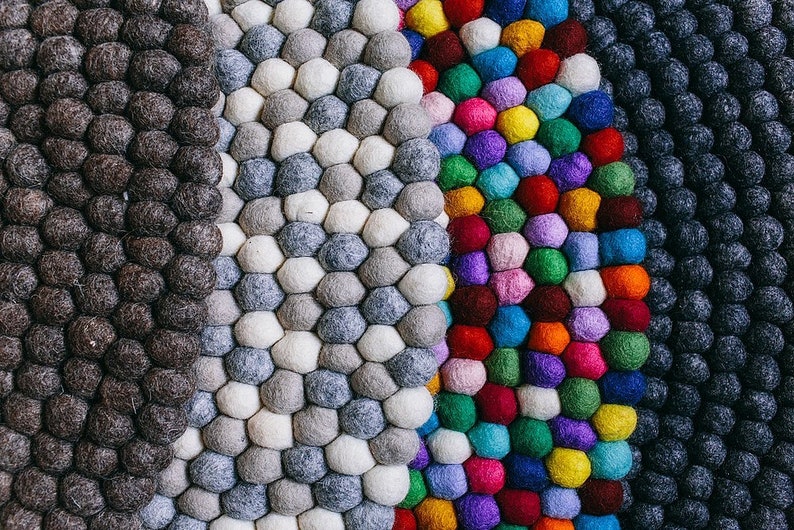 May include: A close-up of a colorful rug made of felt balls. The rug is made up of four sections, each with a different color scheme. The first section is black, the second is gray and white, the third is a rainbow of colors, and the fourth is dark blue.