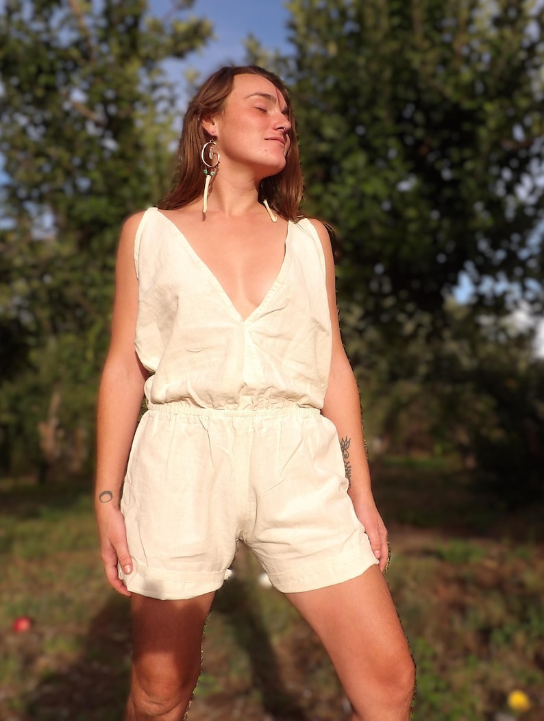 May include: Cream-colored romper with a V-neckline and elastic waist. The sleeveless garment features shorts and is made of a lightweight fabric. The model is wearing hoop earrings with dangling accents. The background is a blurred view of trees.