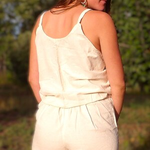 May include: A light beige romper with a scoop neckline and spaghetti straps. The shorts have a relaxed fit. The outfit is made of a lightweight fabric, perfect for warm weather. The model is facing away from the camera.