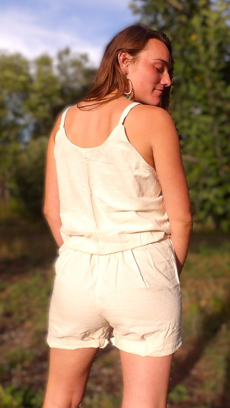 May include: A light beige romper with a scoop neckline and spaghetti straps. The shorts have a relaxed fit. The outfit is made of a lightweight fabric, perfect for warm weather. The model is facing away from the camera.