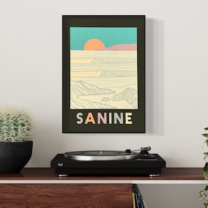 May include: A black framed print featuring a minimalist illustration of a sunset over a beach with the word "SANINE" in colourful letters.