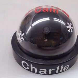 May include: A black dome-shaped security camera replica featuring a dark tinted lens cover. The top is decorated with white snowflake decals and red text reading Santa Cam. The black circular base displays the white printed name Charlie.
