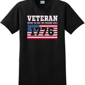 Könnte beinhalten: Schwarzes T-Shirt mit dem Wort "VETERAN" in weißen und lila Blockbuchstaben über einem verblassten Design der amerikanischen Flagge. Der Text "PAYING THE BILL FOR FREEDOM SINCE 1776" ist unter dem Wort "VETERAN" aufgedruckt.