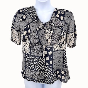 May include: A black and cream-colored patchwork blouse with short sleeves. The blouse features a V-neckline with a ruffled trim. The fabric is patterned with floral and geometric designs. The blouse is displayed on a white mannequin.