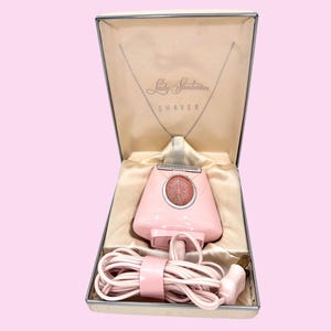 May include: A vintage pink Lady Sunbeam shaver in its original box. The shaver is a light pink color with a circular design on the front. The box is cream-colored with the words "Lady Sunbeam SHAVER" printed on the lid.