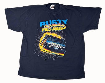May include: Black t-shirt featuring a race car graphic with the text "RUSTY HES BACK! HES BAAD!" in blue and yellow. The graphic shows a race car bursting through a wall of flames. The shirt is a vintage style.